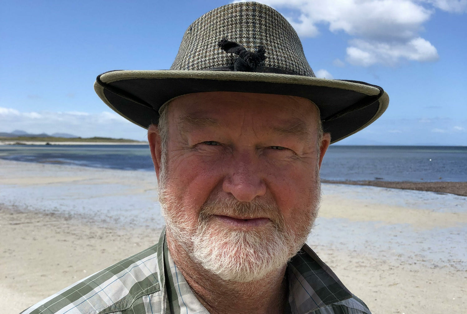 Image of John Kean, he is wearing a hat and has a white beard. The photo is taken by sea with beach and sea in the background.